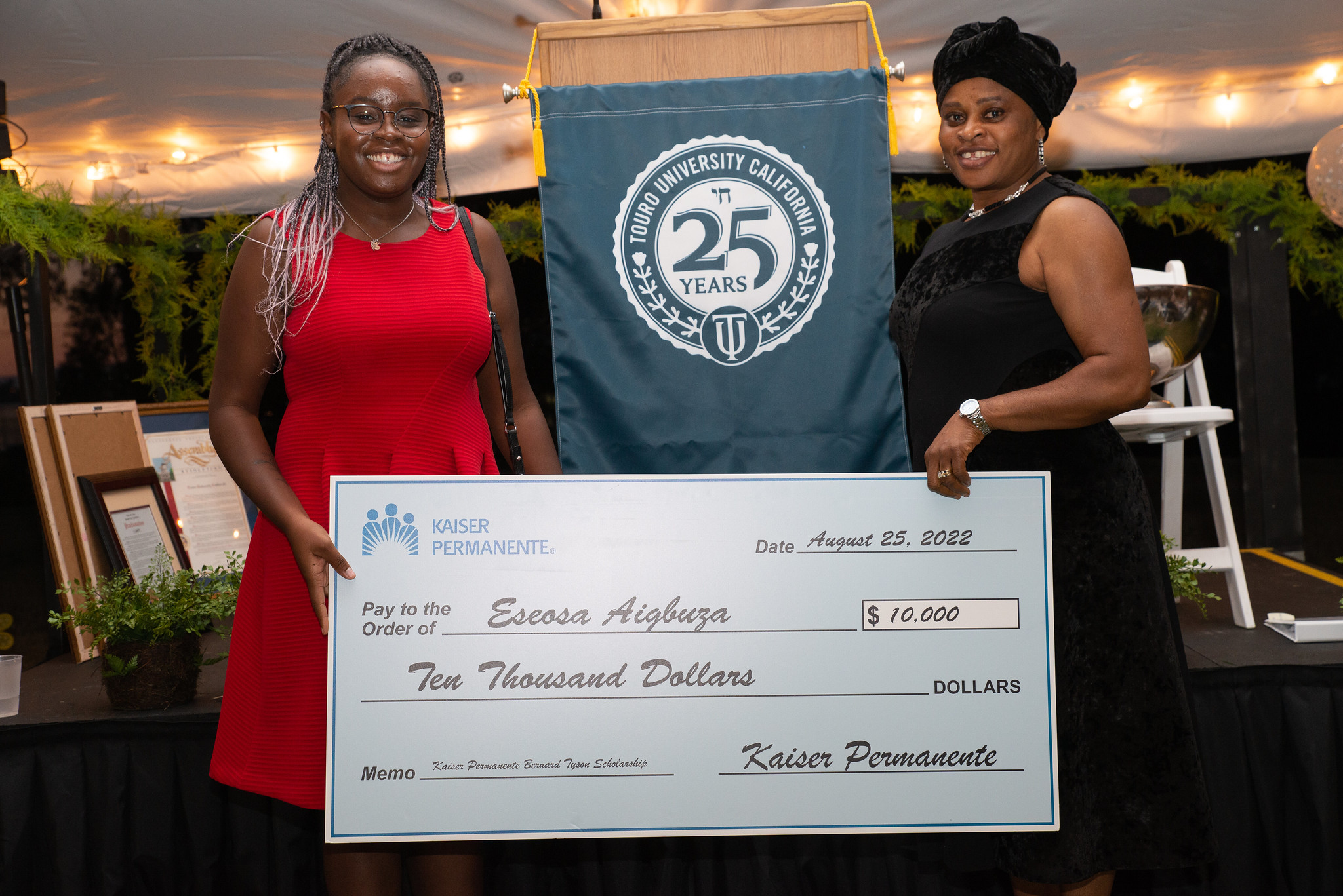 Eseosa “Essie” Aigbuza, a pharmacy student shown standing holding one end of the  Kaiser Permanente Bernard Tyson Scholarship while her mother holds the other