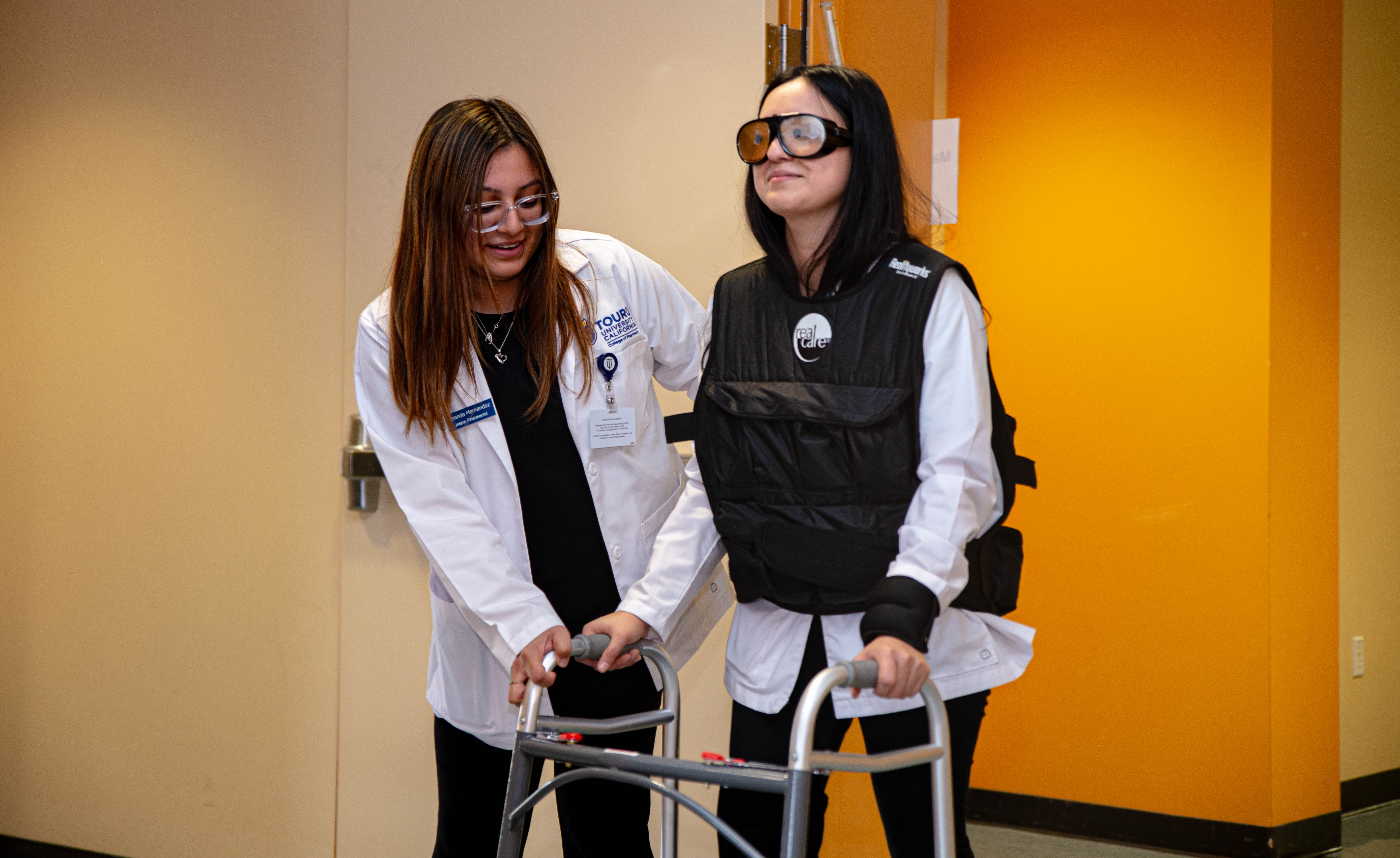 A pharmacy student wearing a weighted vest and glasses that simulate poor vision is guided on a walker by another pharmacy student.