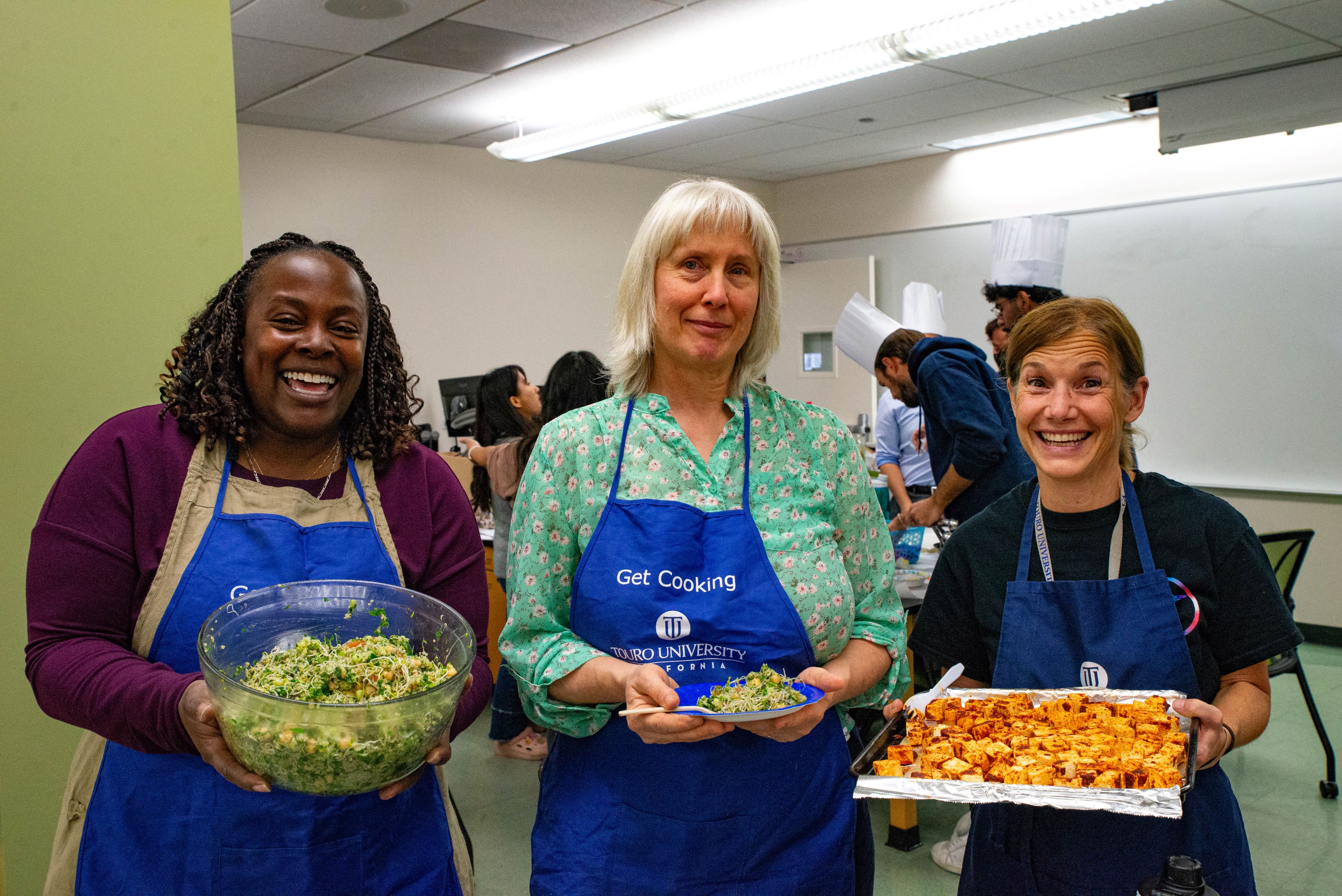 Drs. Grace Marie Jones, Gloria J. Klapstein, and Traci Stevenson from the College of Medicine wearing aprons and holding some of the dishes the medical students cooked up at a pre-clinical nutrition class