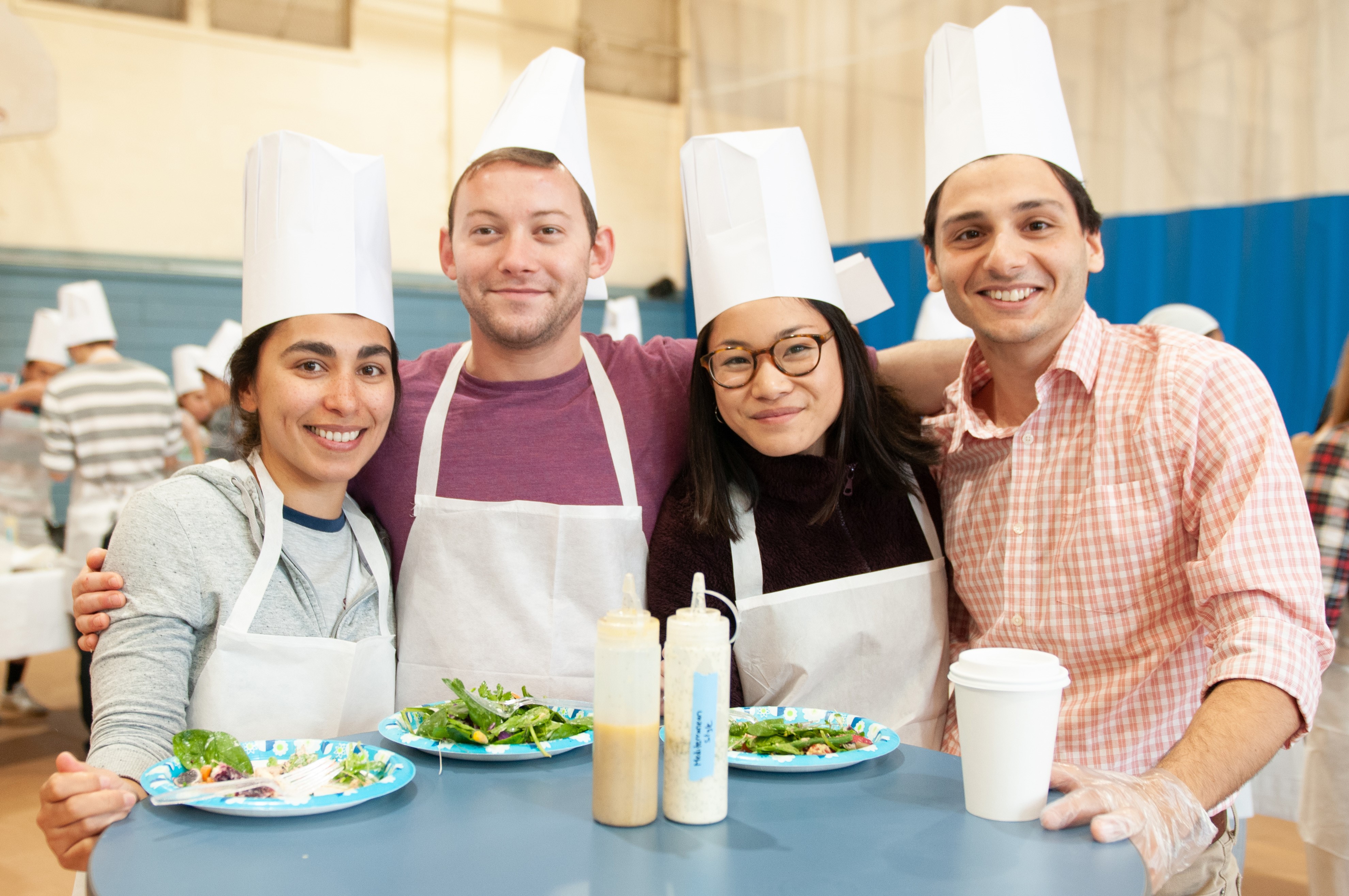 Four student doctors wearing chefs hats stand together eating salads and vegetables on paper plates