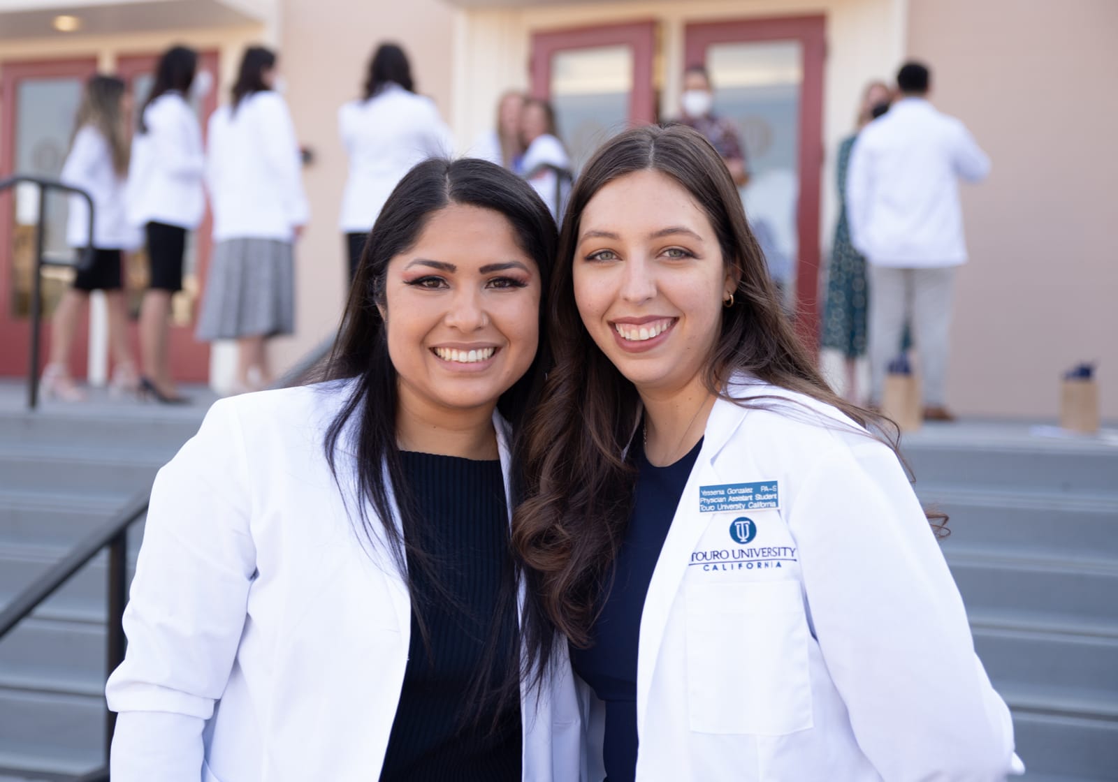 two female PA students in white coats posing outside college building