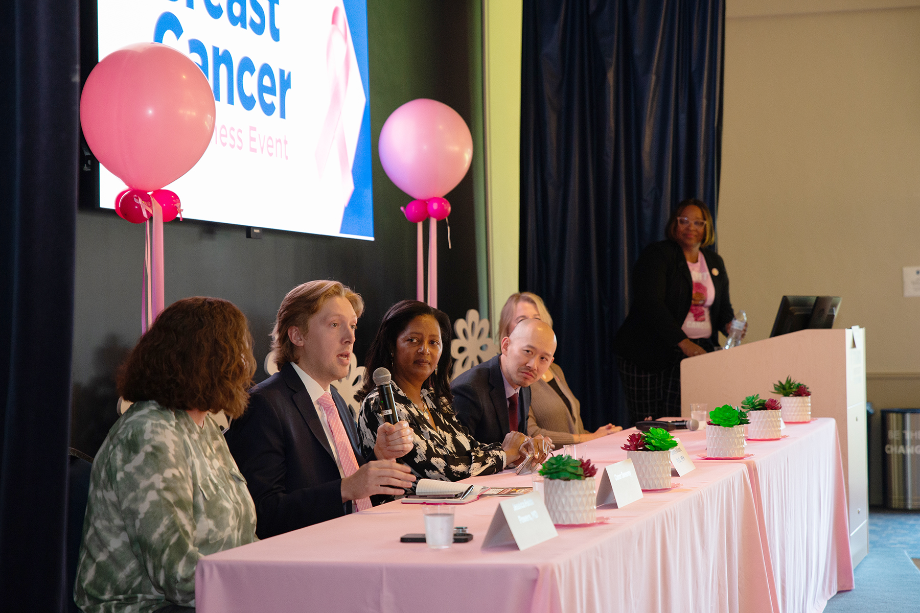 Panelists for the Breast Cancer Awareness event sit on stage behind a table with pink tablecloth in front of a monitor providing information and answering questions at the event.