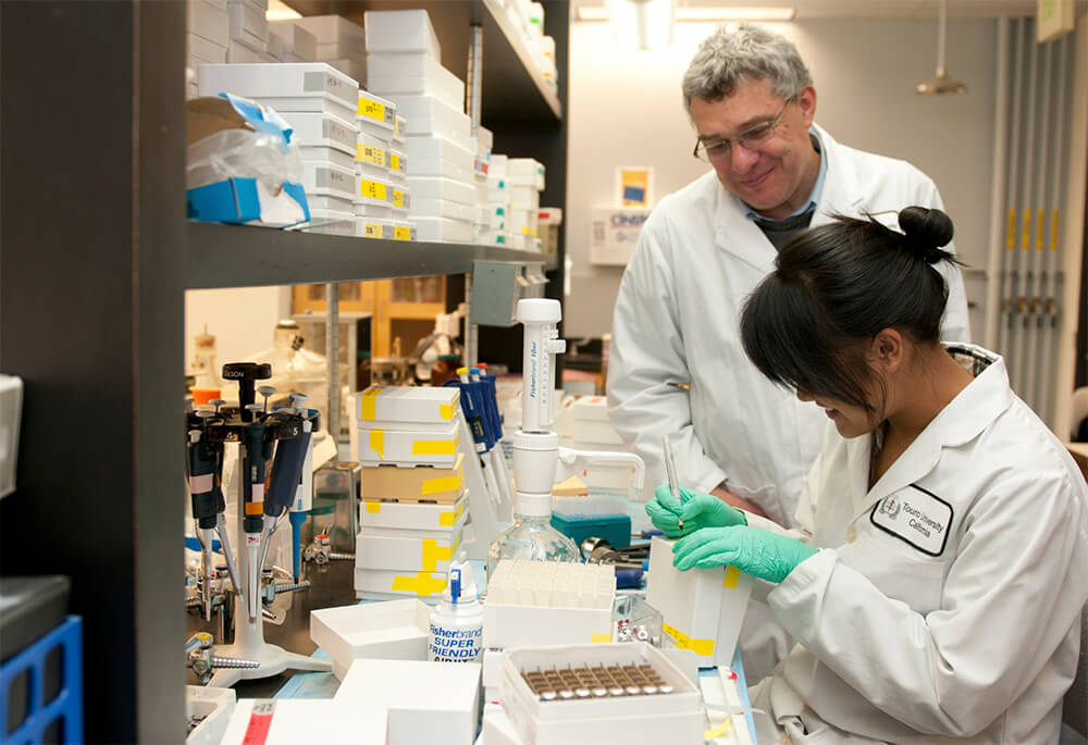 Dr Jean-Marc Schwarz in a lab standing over a female student wearing green latex gloves, watching her write on a box at Touro University California